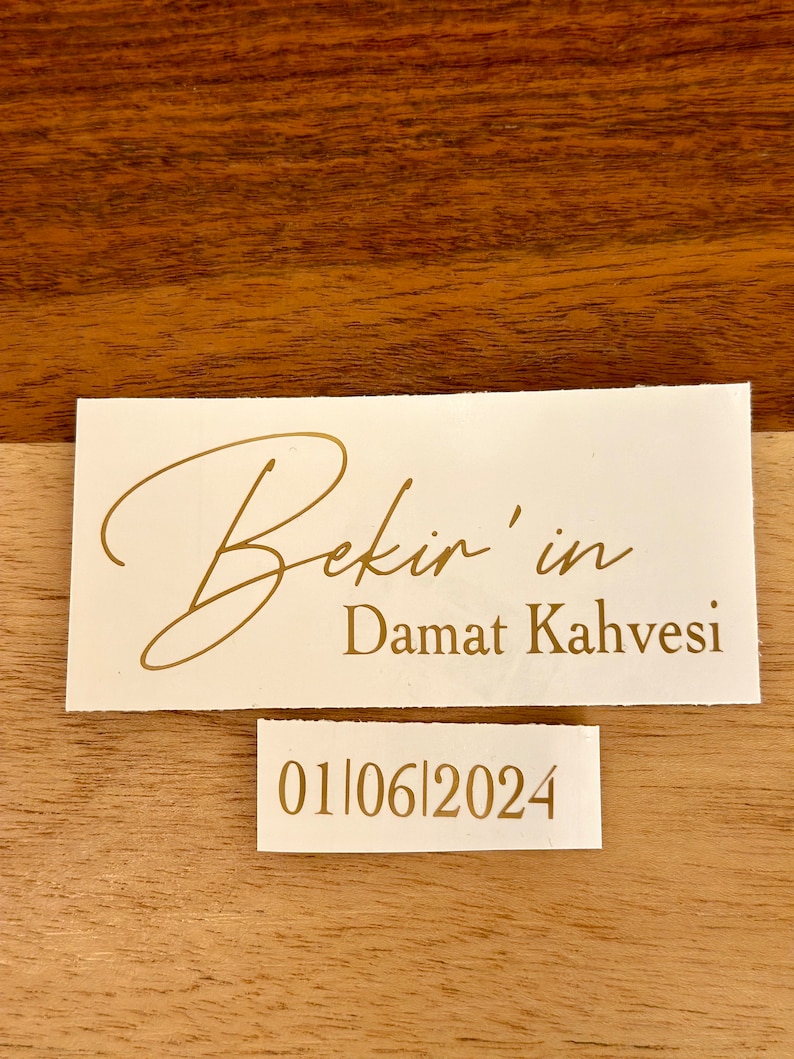 May include: A rectangular white label with gold text reading Bekir'in Damat Kahvesi in a mix of script and serif fonts. Below it, a smaller white label features the gold date 01|06|2024 in a clean serif font. Both labels are placed on a wood surface.