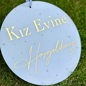 May include: A round, light blue sign with gold lettering and small silver embellishments. The sign has the words "Kız Evine Hoşgeldiniz" in gold script. A light blue ribbon is attached at the top for hanging.