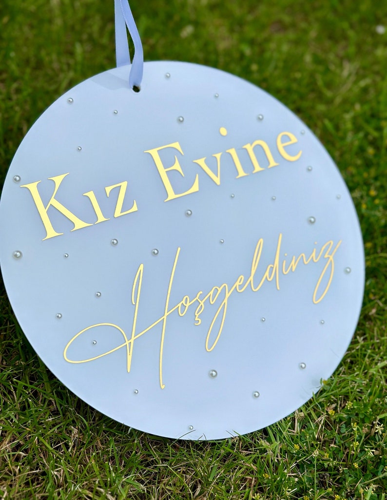 May include: A round, light blue sign with gold lettering and small silver embellishments. The sign has the words "Kız Evine Hoşgeldiniz" in gold script. A light blue ribbon is attached at the top for hanging.