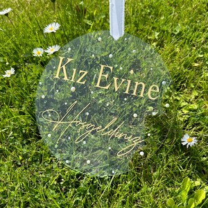 May include: A clear round acrylic sign with gold lettering that reads "Kiz Evine" and "Wedding" in cursive. The sign is hanging from a white ribbon and is surrounded by green grass.