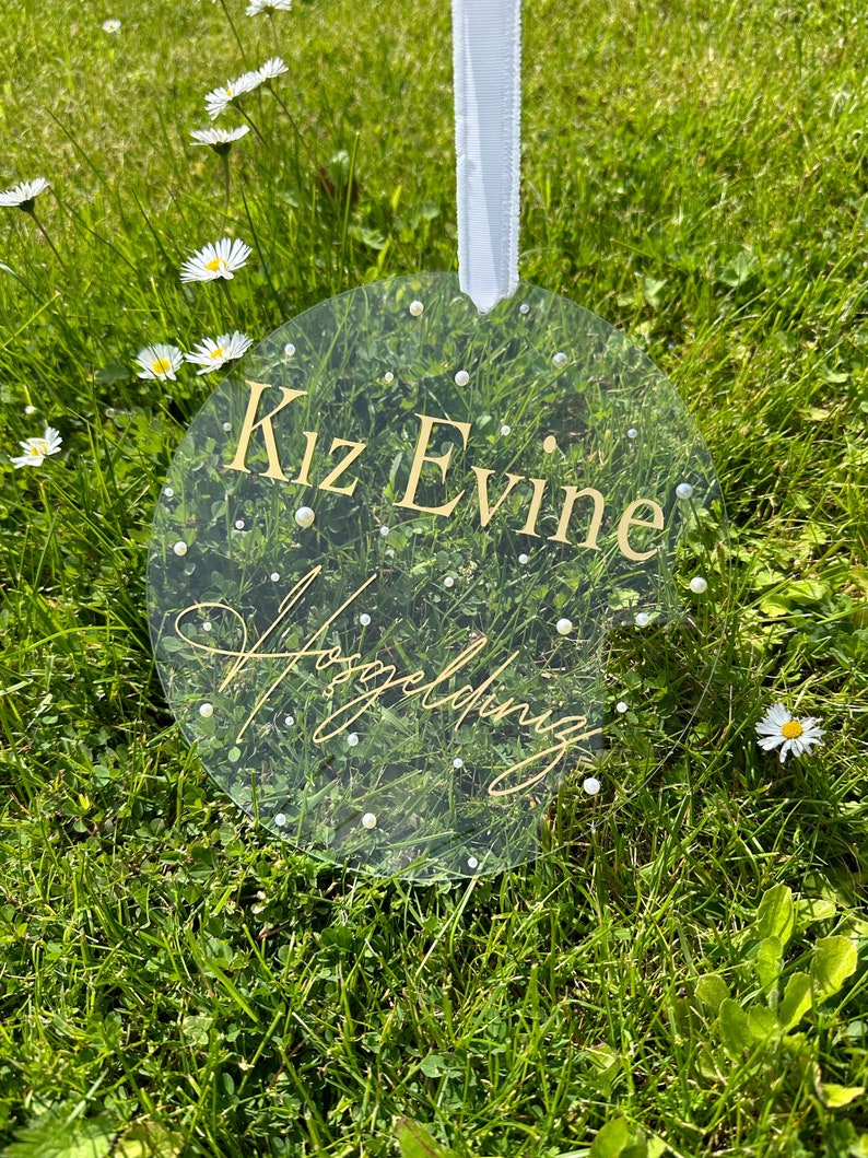 May include: A clear round acrylic sign with gold lettering that reads "Kiz Evine" and "Wedding" in cursive. The sign is hanging from a white ribbon and is surrounded by green grass.