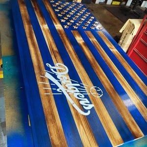 May include: A wooden American flag painted blue with a white "Dodgers" logo and a baseball graphic. The flag is distressed and has a rustic look.