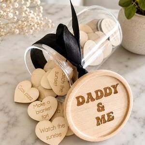 May include: A clear glass jar with a black ribbon tied around the neck, filled with small wooden hearts. The wooden lid is engraved with the text DADDY & ME. Several hearts spill out, featuring engraved phrases like Cook pizza, Make a blanket fort, and Watch the sunrise.