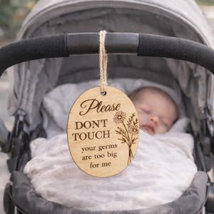 May include: A wooden sign with the text "Please DON'T TOUCH your germs are too big for me" hangs from a baby stroller. The sign has a floral design and is attached with twine. A sleeping baby is visible in the stroller.