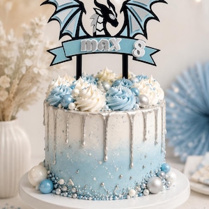 Puede incluir: Un adorno para tarta de varias capas que presenta la silueta de un dragón negro con alas azul claro, situado sobre una pancarta azul claro con el texto max 8 en letras plateadas, sostenido por dos palos negros sobre una tarta glaseada.