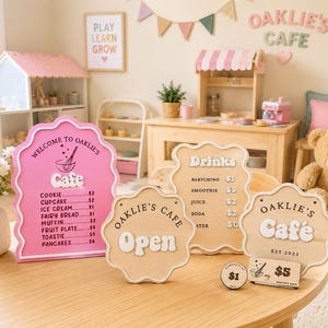 May include: A wooden play cafe set with pink and natural wood signs. The signs display the cafe's name, menu items, and prices. A pink sign says "Welcome to Oaklie's Cafe". Other items include a toy kitchen, a teddy bear, and a sign that says "Open".
