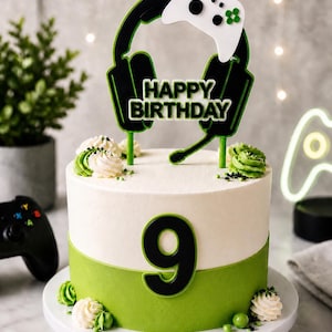 Op de afbeelding: Een verjaardagstaart met twee lagen, witte glazuur en een groene basis. De taart is versierd met een zwart-groene headset en controller taarttopper met de tekst "Happy Birthday". Het nummer "9" staat op de taart.