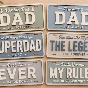 Puede incluir: Seis letreros decorativos rectangulares que se asemejan a placas de matrícula con letras en relieve. Los letreros muestran frases que incluyen World's Best DAD, Legendary DAD, SUPERDAD, THE LEGEND, BEST DAD EVER y DAD'S GARAGE MY RULES.