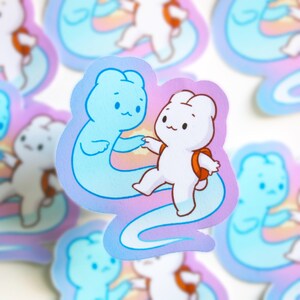 May include: A sticker featuring two cartoon characters. One is a white bear with an orange backpack, reaching out to a blue ghost-like figure. The background has a pastel rainbow gradient. The sticker has a glossy finish.