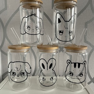 May include: Five clear glass tumblers with bamboo lids and glass straws. Each tumbler has a different black and white cartoon animal face: a dog, cat, bunny, and tiger. The tumblers are designed for cold drinks.
