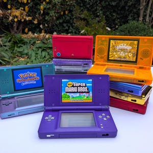 以下が含まれることがあります： 赤、オレンジ、青、紫、ティールなど、さまざまな色のニンテンドーDS携帯ゲーム機コレクション。「New Super Mario Bros.」や「Pokemon Mystery Dungeon」などのゲームが表示されています。