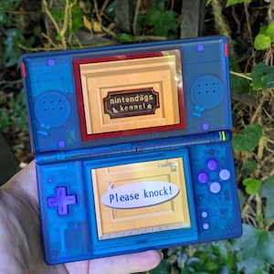 Pode incluir: Uma consola de jogos portátil azul translúcida com botões roxos e ecrã duplo. O ecrã superior mostra um sinal com a inscrição nintendogs kennel e o ecrã inferior mostra uma porta de madeira com um balão de fala que diz Please knock!