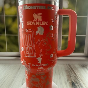 May include: A bright orange Stanley tumbler featuring Monsters, Inc. branding and character graphics in a metallic finish. The cup includes a large side handle, a white lid with a red cap, and text that reads Monsters, Inc., I'm Watching You, and Always Watching.