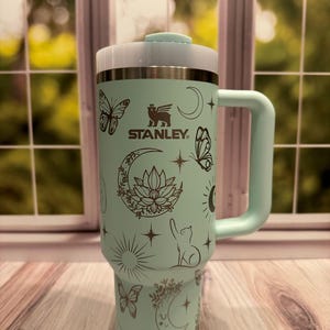 Op de afbeelding: Een lichtgroene Stanley-beker met handvat en deksel, voorzien van donkerbruine lijntekeningen van vlinders, maansikkels, lotusbloemen, zonnen en een kat. Het Stanley-logo is in het donkerbruin bovenaan in het midden gedrukt.