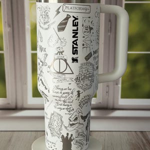 Puede incluir: Un vaso Stanley blanco con asa y tapa negra, que presenta varias ilustraciones de líneas negras y texto relacionado con la serie de Harry Potter, incluyendo el símbolo de las Reliquias de la Muerte, el Sombrero Seleccionador y el texto Platform 9 3/4.
