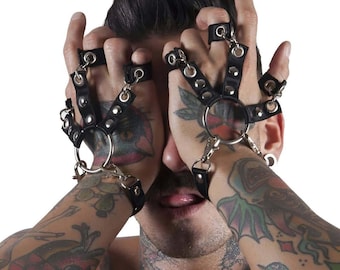 May include: A person wearing black leather fingerless gloves with silver metal rings and studs. The gloves are connected by a chain. The person has tattoos on their arms.