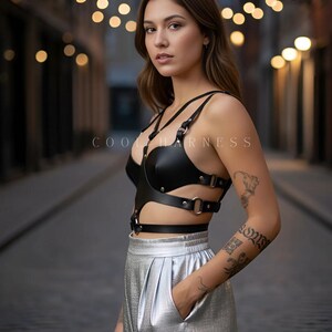 May include: Black leather harness with silver hardware and the text "COOL HARNESS". The harness is worn with silver metallic pleated pants. The image is taken outdoors with blurred background lights.