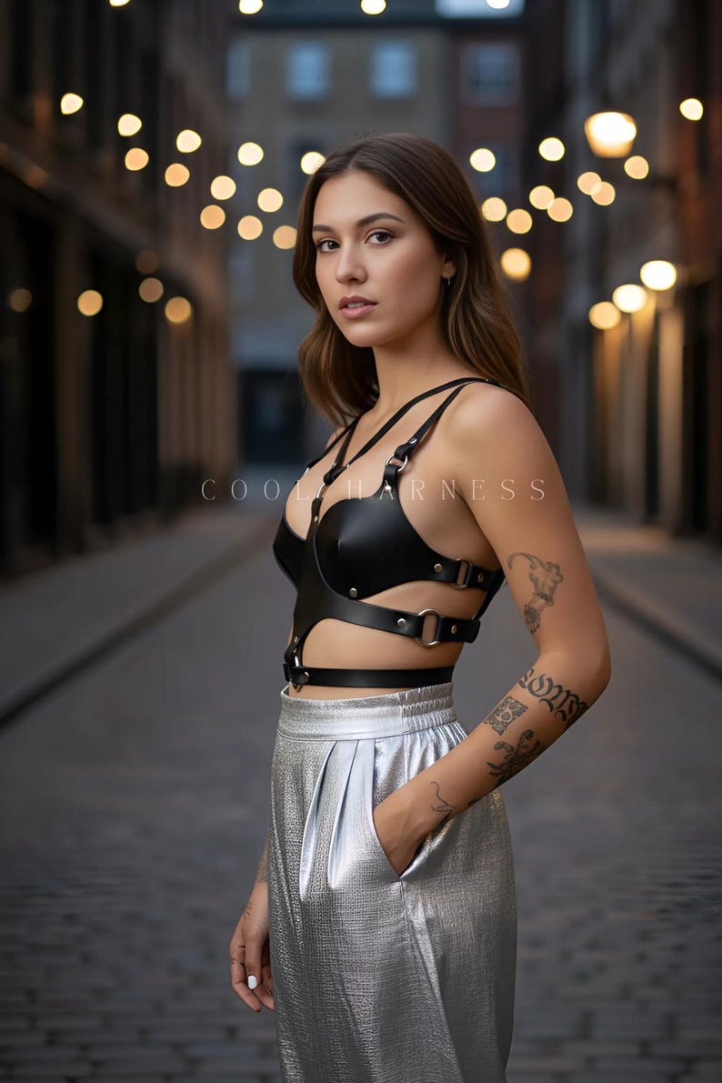 May include: Black leather harness with silver hardware and the text "COOL HARNESS". The harness is worn with silver metallic pleated pants. The image is taken outdoors with blurred background lights.