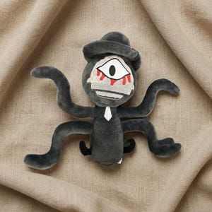 May include: A plush toy in the shape of a creature with one eye, a hat, and multiple arms. The toy is primarily dark gray with white and red accents. It has a small white tie and is set against a neutral background.