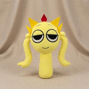 May include: A plush toy in the shape of a yellow creature with a smiling face, black eyes, and yellow hair. The toy has two long, curved arms and a small red gem on top of its head. The background is a neutral beige.