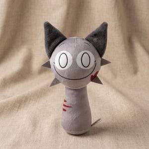 May include: A gray plush toy cat with large, round eyes and a stitched smile. The cat has dark gray ears and spiky accents around its head. Red slash marks are on the neck. The toy is on a beige fabric background.