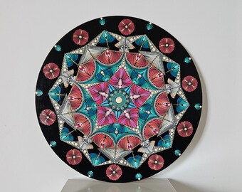 May include: A round mandala painted on a black background with a colorful design of pink, blue, and white. The mandala is surrounded by a ring of small red flowers.