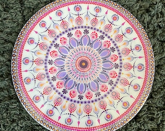 May include: A white circular mandala with a pink, purple, and gold dot pattern. The mandala is painted on a round wooden surface.