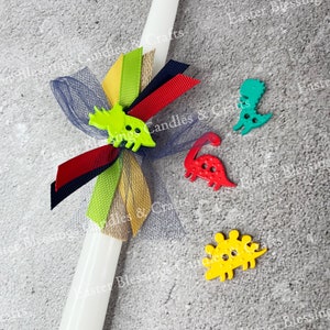 May include: A white candle decorated with a blue tulle bow, red, black, and yellow ribbons, and green, red, and yellow dinosaur-shaped buttons. The candle is on a gray textured surface.