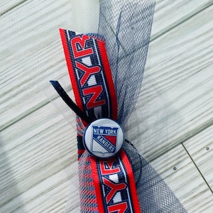 Può includere: Una candela bianca con un nastro blu e rosso con le parole "NY Rangers" su di esso. Un bottone blu e bianco con il logo dei New York Rangers è attaccato al nastro.