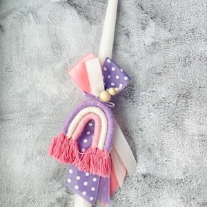 May include: A white candle with a pink and purple rainbow-shaped decoration, white and pink ribbons, and white polka dot ribbon.