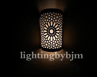May include: A black metal wall sconce with a geometric pattern and a white light shining through it. The sconce is lit and casting a warm glow on the wall. The text "lightingbybjm" is visible below the sconce.