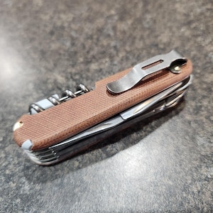 Op de afbeelding: Een bruin multifunctioneel zakmes met een zilveren clip en een kurkentrekker. Het mes heeft een bruin, getextureerd handvat.