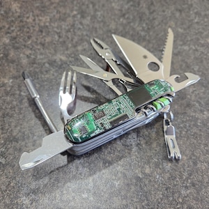 May include: A multi-tool with a green circuit board pattern on the handle. The tool features several extended stainless steel implements, including a knife blade, pliers, scissors, a fork, a screwdriver, and a bottle opener, all attached to a central frame.