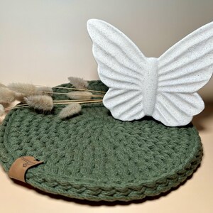 Könnte beinhalten: Ein runder, grüner Häkeluntersetzer mit einem weißen Keramik-Schmetterling darauf. Der Untersetzer hat ein braunes Leder-Etikett mit dem Wort "Made" darauf.