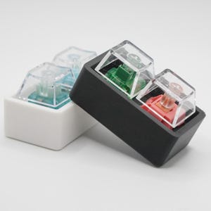 May include: Three clear plastic mechanical keyboard keycaps with different colored switches. The keycaps are in a black and white plastic case.