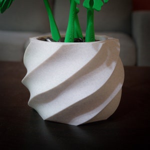 May include: A white, textured, spiral-shaped planter with green plant stems and leaves. The planter has a unique, modern design, and is filled with small, dark stones. The plant stems and leaves are a bright green color.