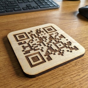 May include: Square wooden coaster with a laser-etched QR code. The coaster is made of light-colored wood with a dark brown etched design. The coaster is on a wooden desk next to a keyboard and mouse.