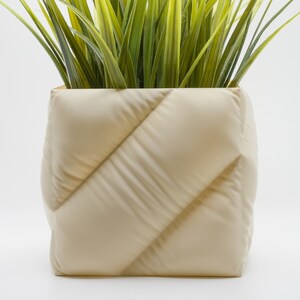 May include: A cream-colored planter with a quilted design. The planter is filled with green grass-like plants.