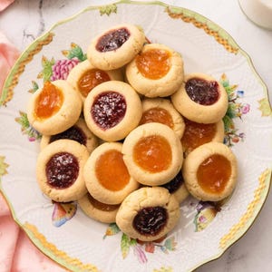 Könnte beinhalten: Ein Teller mit Daumendruck-Keksen mit verschiedenen Fruchtkonfitüren. Die Kekse sind goldbraun mit einer kreisförmigen Vertiefung, die mit roter oder orangener Marmelade gefüllt ist. Der Teller hat ein Blumendesign mit grün-goldener Verzierung.