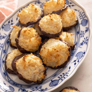 May include: A serving plate with a blue and white pattern displays several coconut macaroons dipped in dark chocolate. The macaroons have a golden-brown, toasted exterior and a soft, white interior. The plate has a delicate floral design.