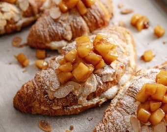 Peut inclure: Gros plan sur plusieurs croissants dorés garnis de dés de pommes et saupoudrés de sucre glace. Des amandes effilées sont visibles. Les pâtisseries sont disposées sur une plaque de cuisson recouverte de papier sulfurisé.