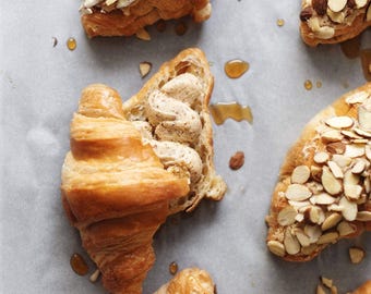 Peut inclure: Vue aérienne de plusieurs croissants dorés, certains garnis d'amandes effilées. Un croissant est coupé, révélant une garniture crémeuse. Des filets de sirop sont dispersés autour des pâtisseries sur une surface gris clair.