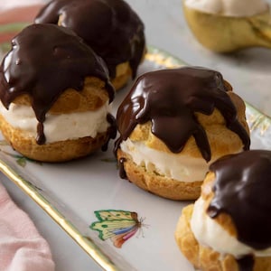 Pode incluir: Vários profiteroles com cobertura de ganache de chocolate preto são apresentados numa travessa decorativa. Os profiteroles são recheados com creme. Uma colher dourada contém uma porção de creme. A travessa tem um desenho de borboleta.