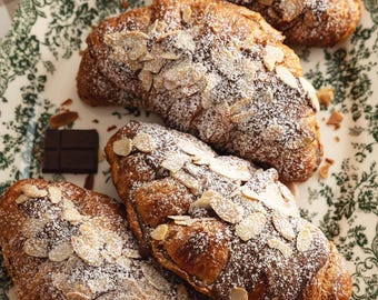 Peut inclure: Quatre croissants aux amandes saupoudrés de sucre glace sur une assiette décorative. Les croissants sont dorés avec des amandes effilées sur le dessus. Un petit morceau de chocolat noir est posé sur l'assiette. L'assiette a un motif floral vert et blanc.