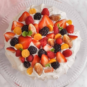 Może przedstawiać: Deser Pavlova na przezroczystym szklanym talerzu, posypany świeżymi truskawkami, malinami, jeżynami, figami i miechunką. Podstawa z bezy pokryta jest bitą śmietaną, tworząc kolorową i apetyczną prezentację.