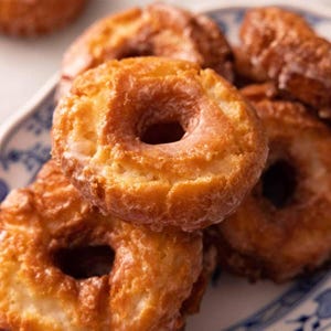 May include: A close-up shot of several glazed donuts on a blue and white patterned plate. The donuts are golden brown with a glossy glaze. The donuts are stacked on top of each other, with some visible in the background.