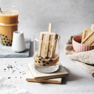 May include: A refreshing beverage with a unique presentation. A glass of iced tea with boba pearls and a popsicle. The popsicle is in the shape of a rectangle with vertical lines. Other glasses and a bowl with popsicles are also visible.