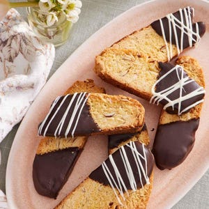 May include: A light pink oval plate holds several biscotti cookies. The biscotti are dipped in dark chocolate and drizzled with white chocolate. The cookies are arranged on a light green tablecloth, with a small vase of flowers in the background.