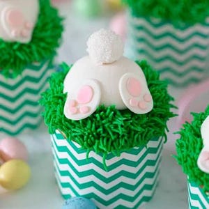 May include: Easter-themed cupcakes with white bunny bottoms and green frosting resembling grass. The cupcakes are in chevron-patterned liners, and pastel-colored candies are scattered around them. A festive and delicious treat.
