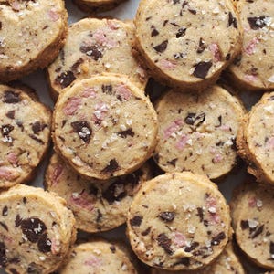 Può includere: Primo piano di diversi biscotti rotondi con pezzi di cioccolato e pezzi rosa, cosparsi di sale grosso. I biscotti sono marrone chiaro e disposti vicini.